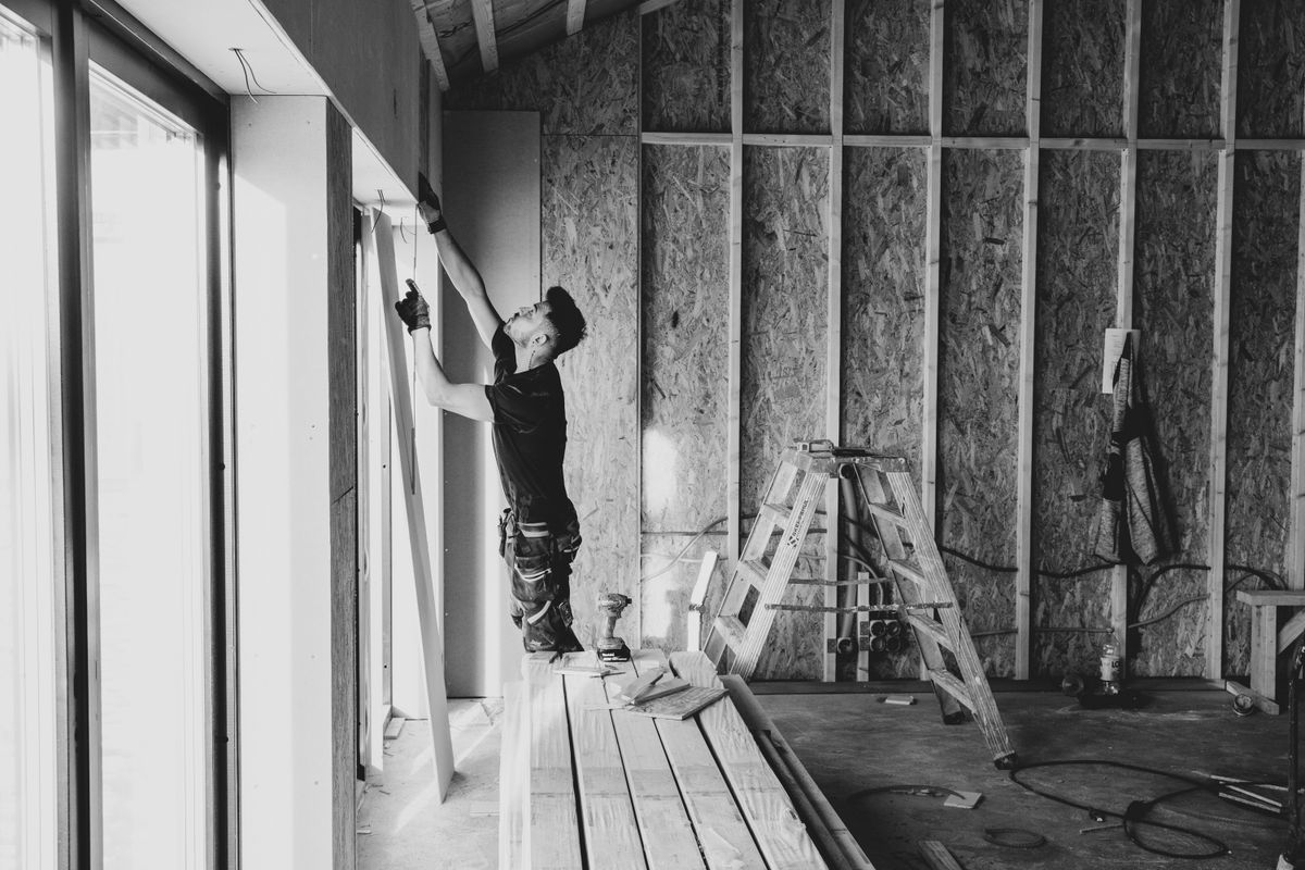 Handwerker hebt in einem Rohbauraum mit Holzständerwerk eine Platte an eine Fensterlaibung (Symbolfoto)