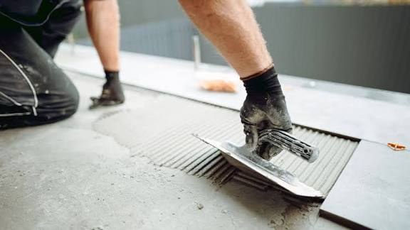 Handwerker mit Arbeitshandschuh trägt mit einer Zahnkelle Kleber auf einen Betonboden auf, daneben liegt eine Platte (Symbolfoto)